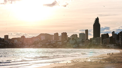 Sea by modern buildings against sky during sunset