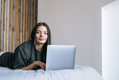 Beautiful smiling woman freelancer with dark long hair in casual using laptop sitting on bed at home