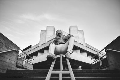 Low angle view of young man sitting on staircase railing against sky