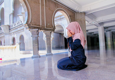 Full length of woman in hijab holding counting rosary beads while praying at mosque