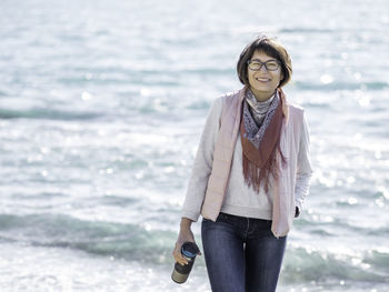 Smiling woman walking on seaside with travel mug. vacation on ocean coast. thermos with hot beverage