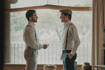 Side view of handsome guys in shirts and trousers with suspenders talking near seat with composition of blooms and window in room