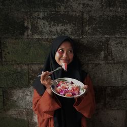 Woman wearing hijab eating food against wall