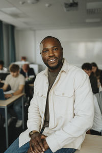 Portrait of smiling male teacher sitting on desk in classroom at high school