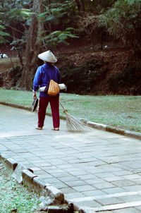 Rear view of man holding broom and walking on footpath at park