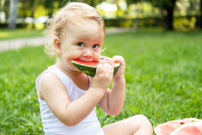 Funny smiling kid boy in white bodysuit eating watermelon at green lawn