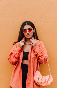 Confident young female millennial wearing trendy outfit and modern sunglasses standing with bag against plaster orange wall