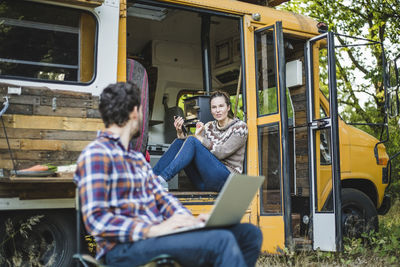 Woman sitting at entrance of caravan while talking with friend using laptop during camping in forest