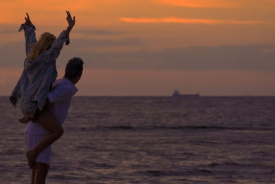 Man piggybacking woman while standing at beach against sky during sunset