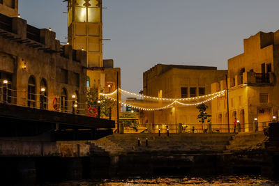 Illuminated bridge over river by buildings against sky at night