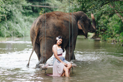 Woman sitting by elephant on swing in river at forest