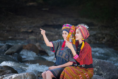 Smiling women wearing traditional clothing while sitting on rocks by stream