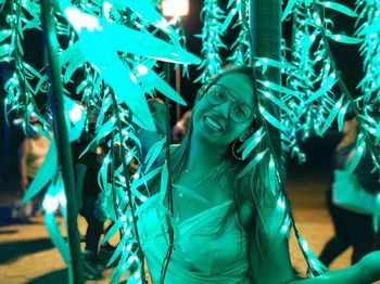 Portrait of young woman smiling while standing amidst turquoise lights at night