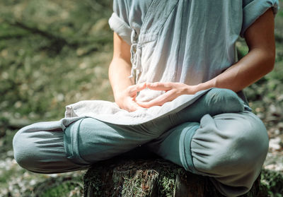 Cropped unrecognizable man in traditional clothes sitting on rock in lotus pose and meditating during kung fu training in forest