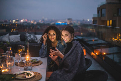 Smiling woman showing smart phone to female friend during social gathering on building terrace