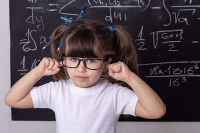 Portrait of cute girl with hands covering ears standing against blackboard
