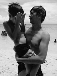 Shirtless man carrying son while standing at beach against sky