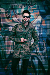 Portrait of handsome young man wearing sunglasses while standing against graffiti on shutter