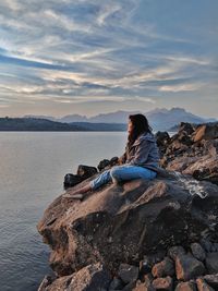 Rear view of man sitting on rock looking at shore against sky