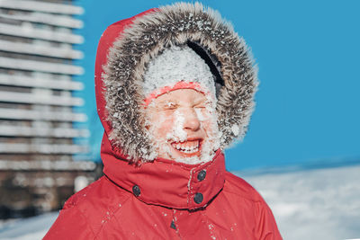 Funny cute adorable caucasian smiling girl in red jacket parka squinting eyes from bright sun. 