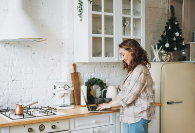 Attractive smiling woman with curly hair in plaid shirt with laptop near window at bright kitchen 