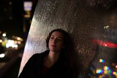 Woman looking away while standing by metal column at night