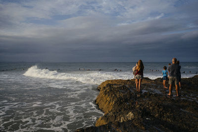 Family standing on rock at sea shore against cloudy sky