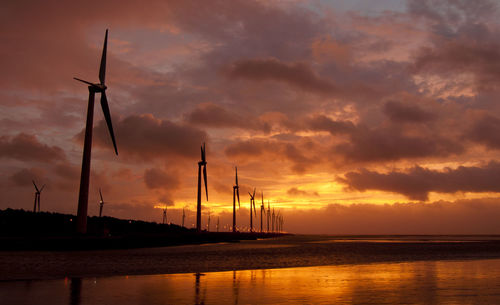 Silhouette wind turbines by sea against sky during sunset