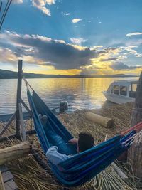 Man relaxing on hammock by sea against sky during sunset