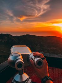 Cropped hand of man holding coin-operated binoculars against sky during sunset