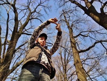 Low angle view of man gesturing finger frame while standing by bare trees
