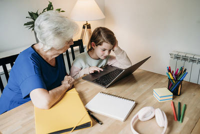 Senior woman looking at granddaughter e-learning on laptop at table