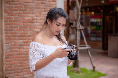 Young woman holding camera while standing by brick wall