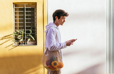 Side view of young male standing on city street with eco friendly mesh bag with groceries and using smartphone