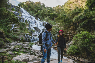 Smiling woman with friend standing by waterfall in forest