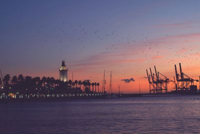 Sea by illuminated building against sky during sunset