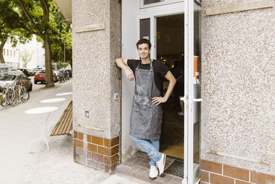 Smiling male cafe owner with hand on hip leaning on wall at doorway