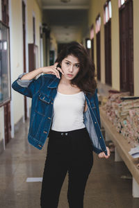 Portrait of young woman holding denim jacket while standing in corridor