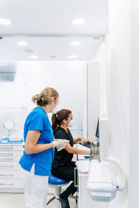 Side view of nurse in medical clothes standing against female dentist looking at screen while taking x ray picture