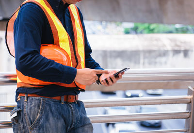 Midsection of man holding hardhat while using mobile phone on railing