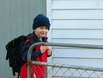 Portrait of smiling boy carrying backpack standing against wall