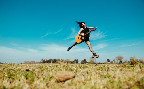 Woman with guitar jumping over field against sky