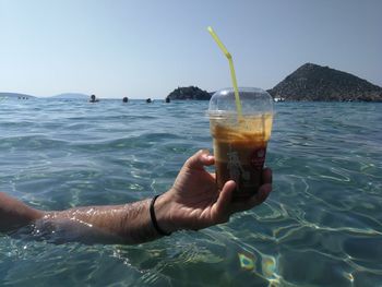 Hand holding drink by sea against sky