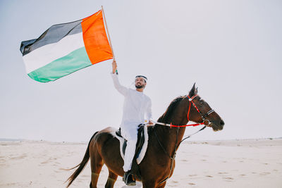 Man holding flag while horseback riding against sky