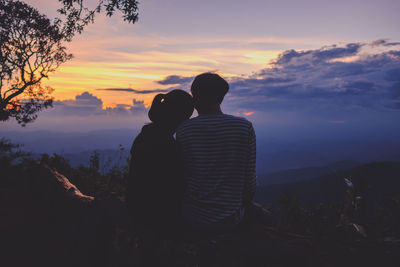 Rear view of silhouette couple sitting on mountain against sky during sunset