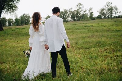 Rear view of wedding couple holding hands while walking on grassy field