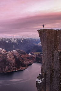 Man on cliff by fjord against sky during sunset