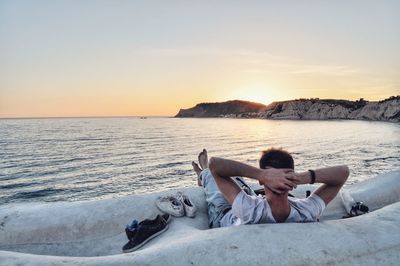 Man lying down by sea against sky during sunset