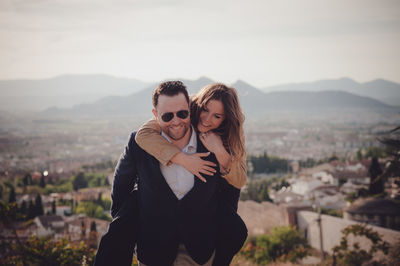 Man giving piggy back ride to happy woman standing against cityscape