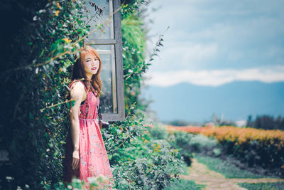 Portrait of woman standing by window against plants in back yard